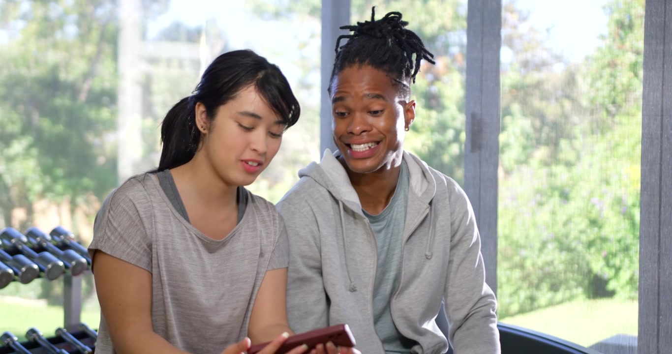 African American man and Asian woman on bench, woman showing red smartphone, agreeing and smiling