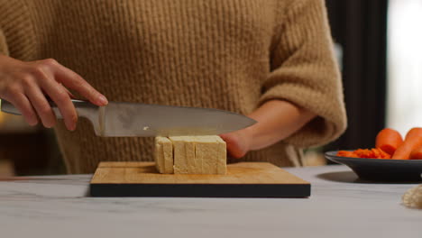 close up of woman at home in kitchen preparing healthy vegetarian or vegan meal slicing tofu on board with knife 2