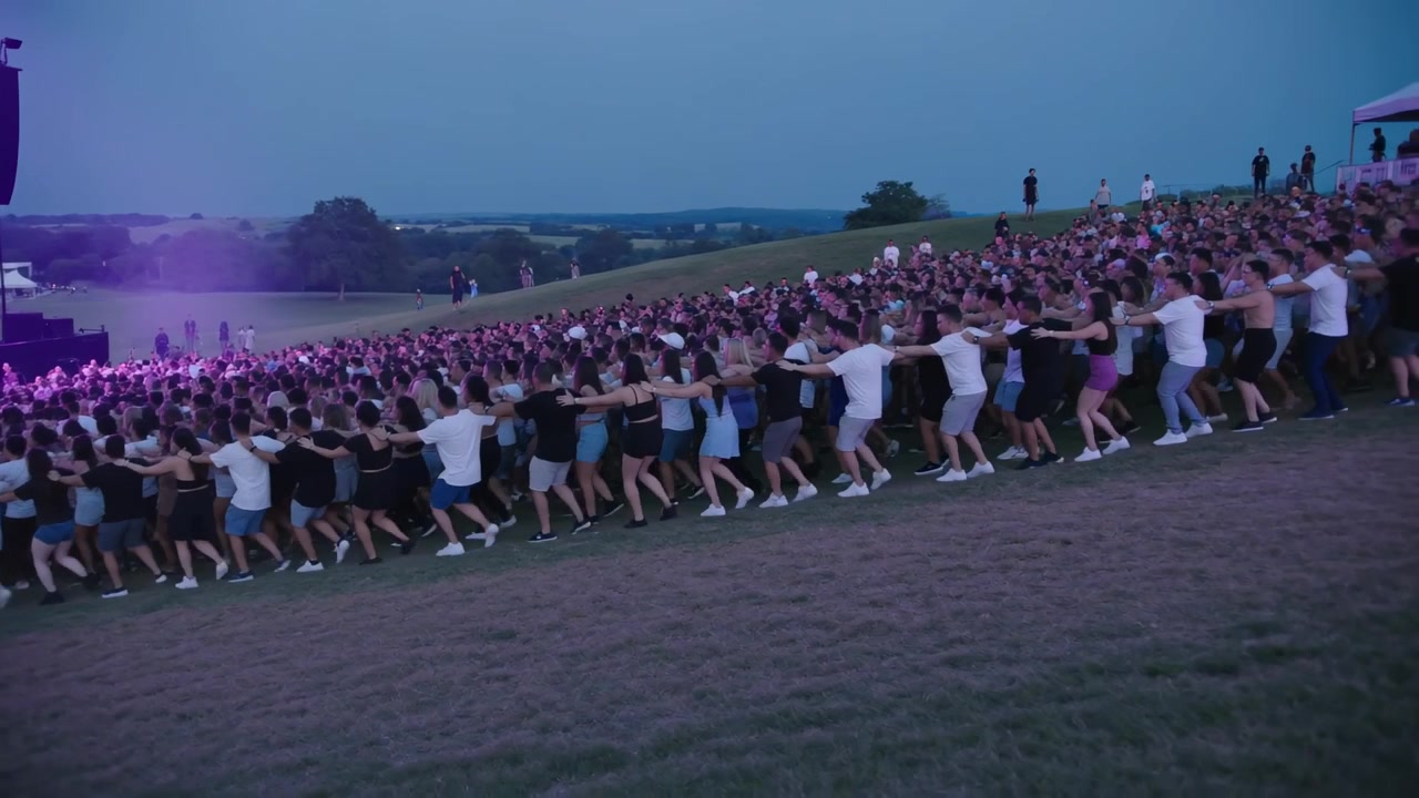 Une foule unie en t-shirts et shorts marchant sur une colline herbeuse au crépuscule, réagissant à la musique de la scène.