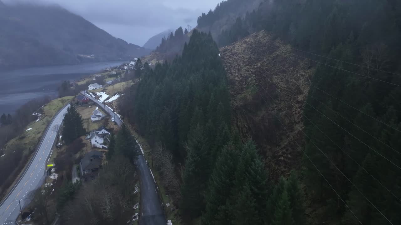 Drone segue linee elettriche ad alta tensione con il fiordo di Sorfjorden sulla sinistra e la strada E16 a Romslo in Norvegia. Infrastruttura elettrica con tempo nebbioso e piovoso.