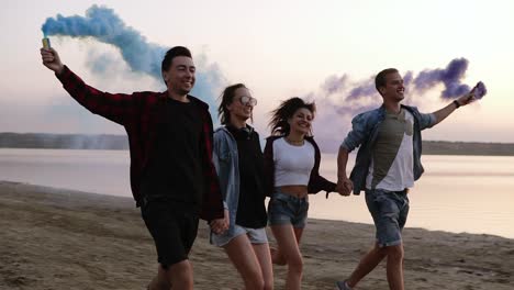 Young-Four-People-Running-With-Colored-Smoke-Grenade-In-Their-Hands-By-The-Sea-During-Sunset