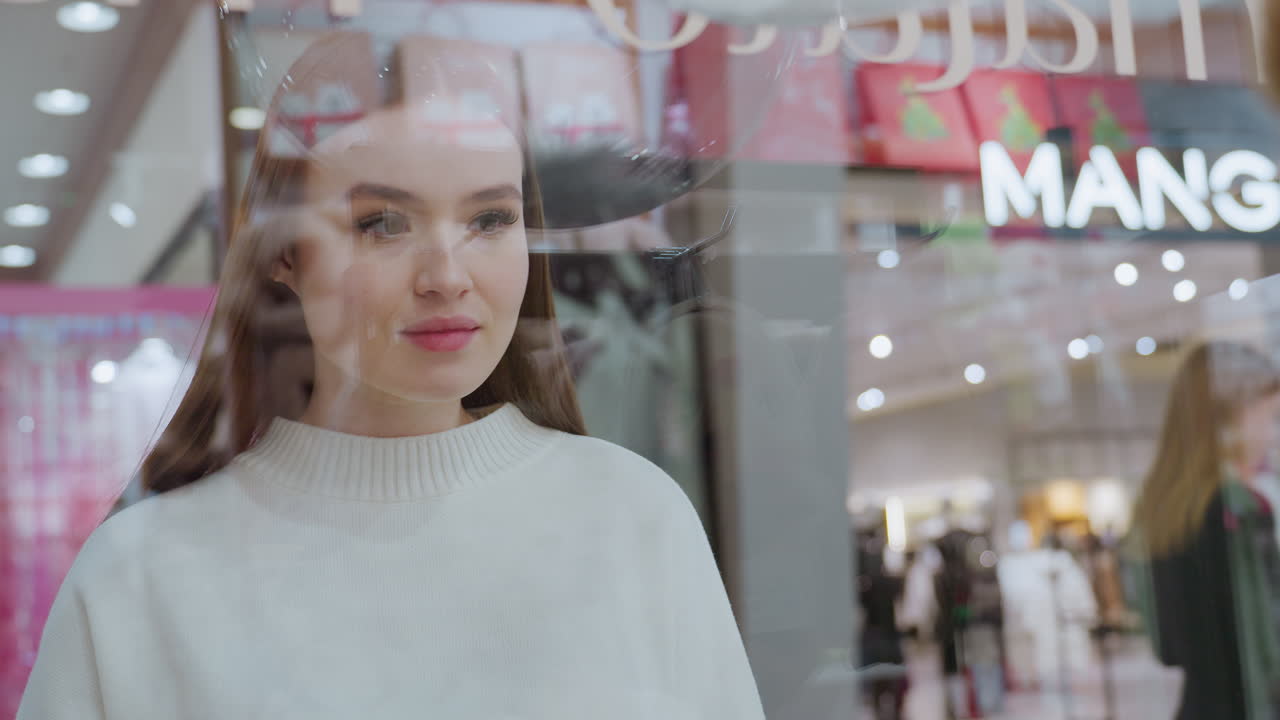 Woman standing in front of a glass display, observing something in the background of a mall, reflections on the glass, with people passing by