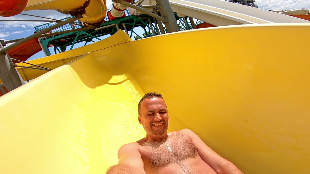 Joyful male going down from yellow pipe outdoors. Man having fun and sliding down in a water slide in summer at a sunny day.