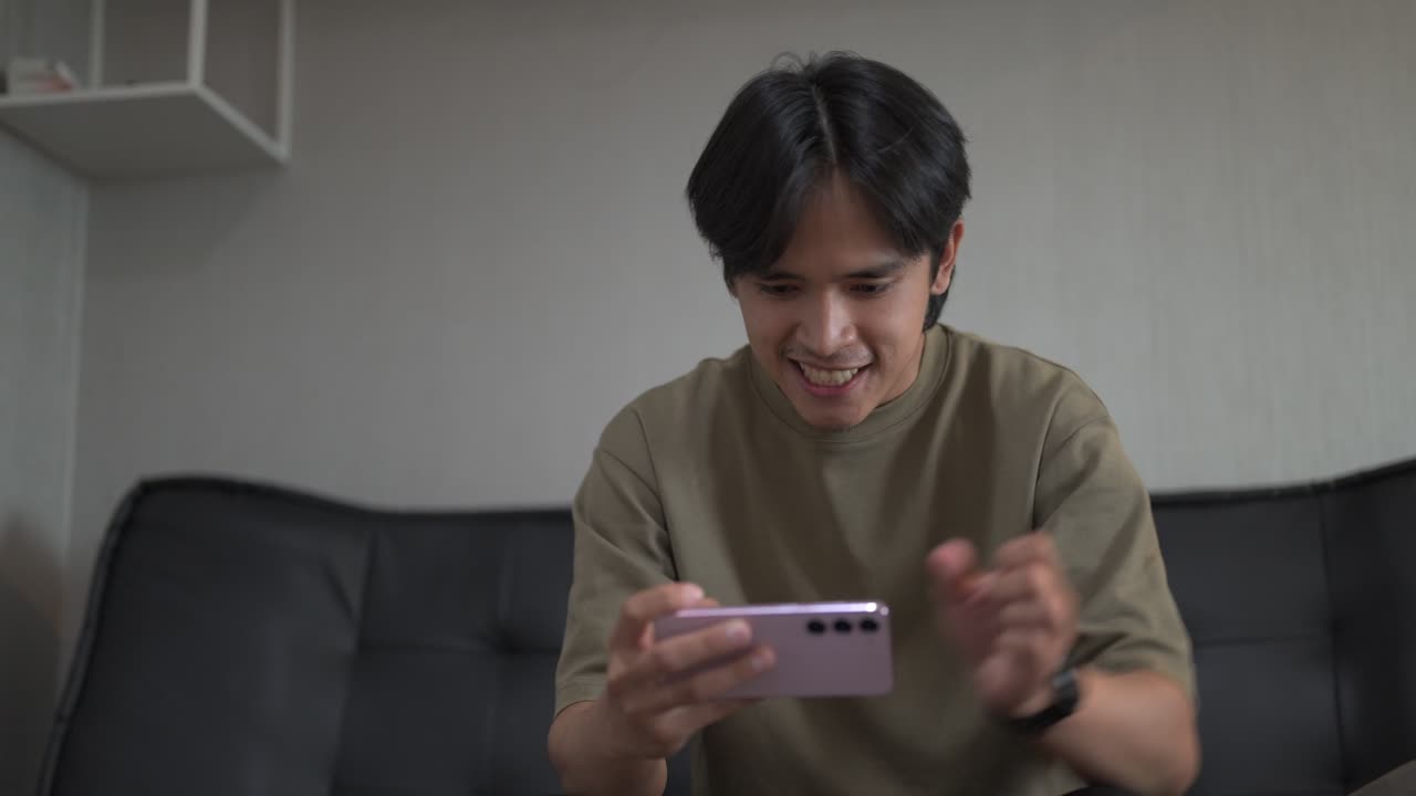 Handsome young Asian Thai man sitting at sofa indoors while playing games with mobile phone