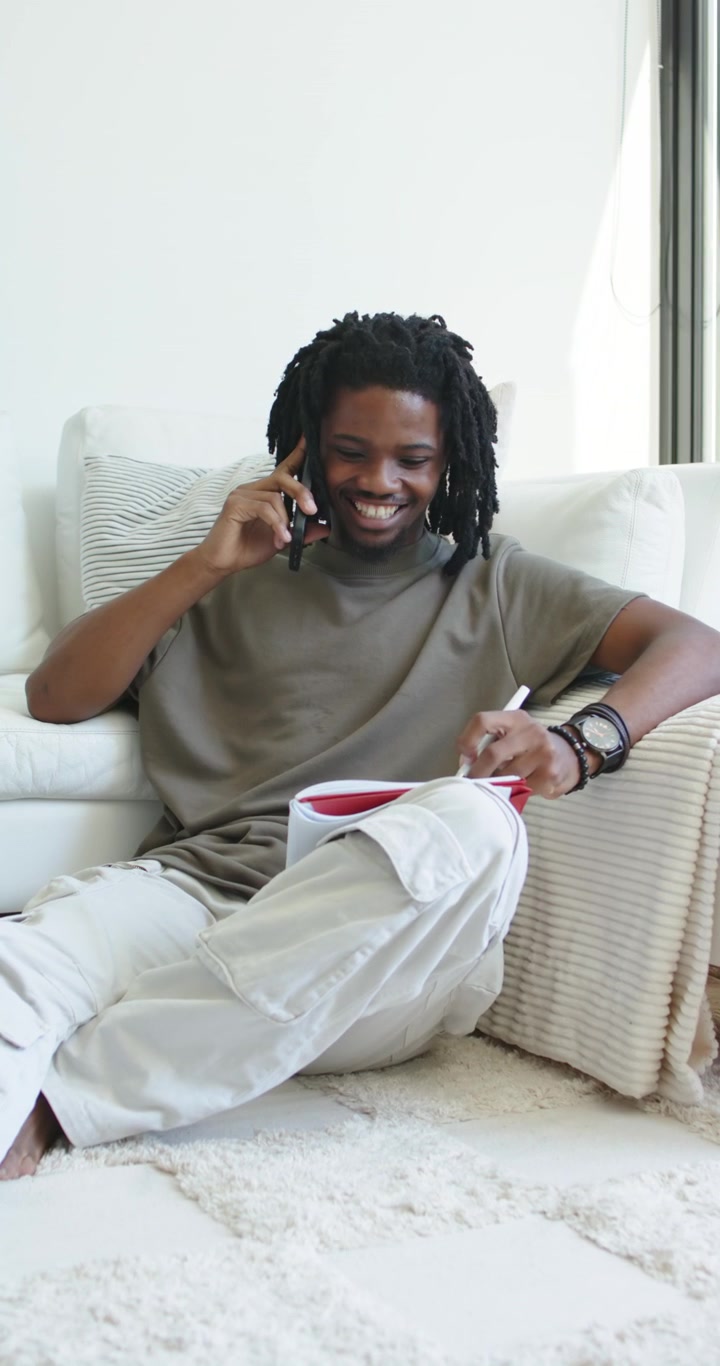 Video verticale: Uomo afroamericano che risponde a una chiamata sullo smartphone mentre prende appunti su un quaderno rosso a casa