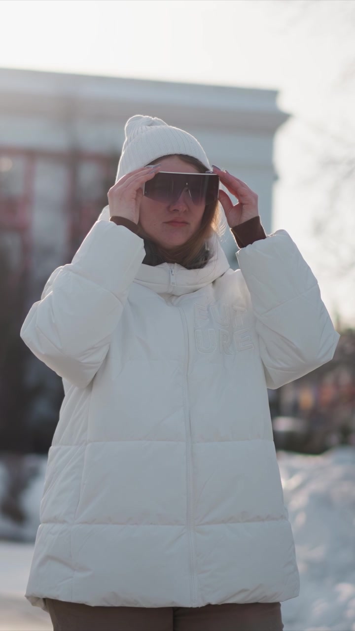 Woman wearing white puffer jacket and beanie holds sunglasses in outdoor winter setting adjusting them while standing on snow with buildings and trees in background. Woman wears headphones