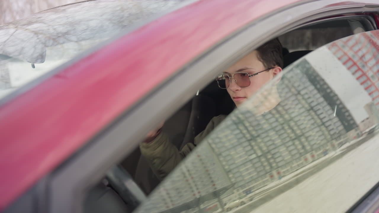 student dressed in winter jacket sitting in parked car rolls up window while scratching head with faint winter landscape reflected on glass creating calm seasonal mood and interior contrast