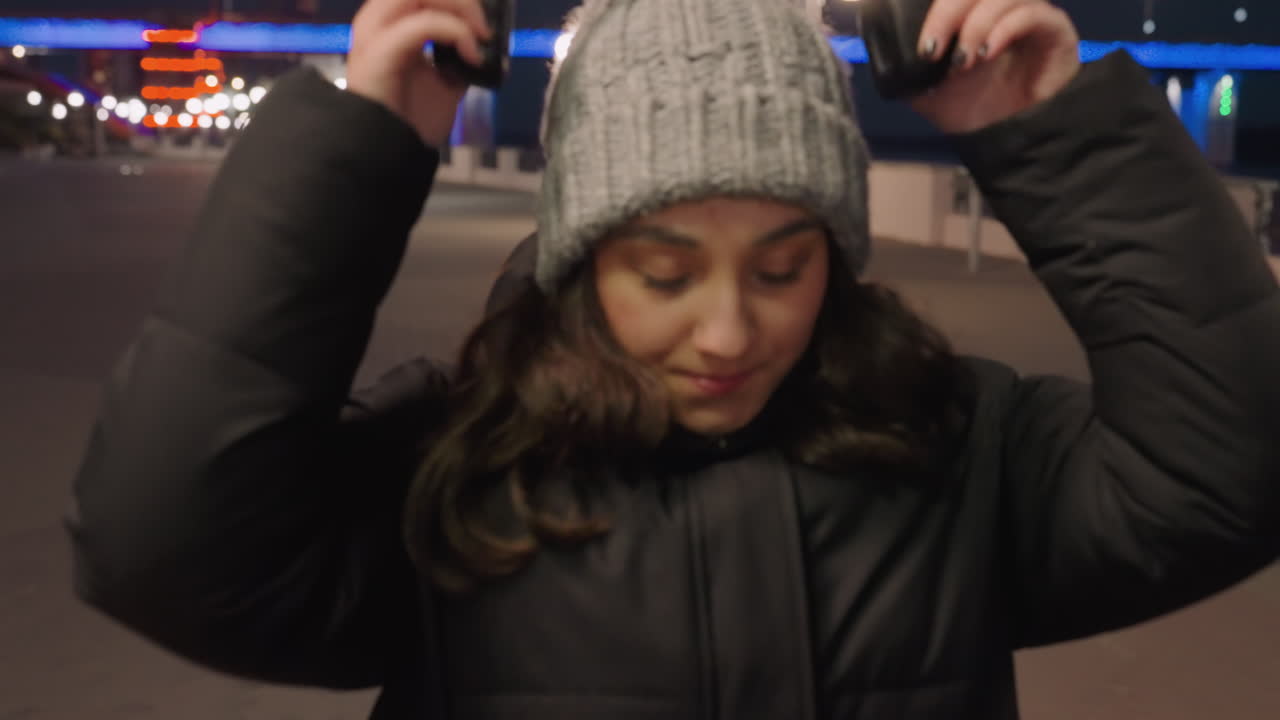 Mulher à beira-rio à noite com fones de ouvido em calçadão vazio, gorro e casaco acolchoado, luzes suaves e azuis da ponte, balançar lento da cabeça ao ritmo da música, humor reflexivo, bokeh cinematográfico, frio de inverno.