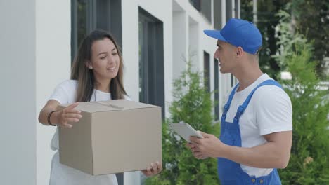 beautiful woman meets a delivery man who gives her a parcel box beside her home, courier checks information on tablet. home delivery, online shopping