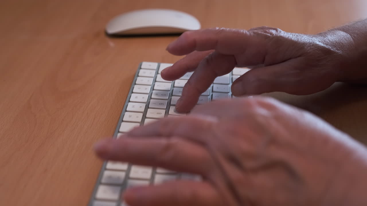 händerna på en äldre kvinna som skriver på ett vitt datortangentbord på ett kontorsbord