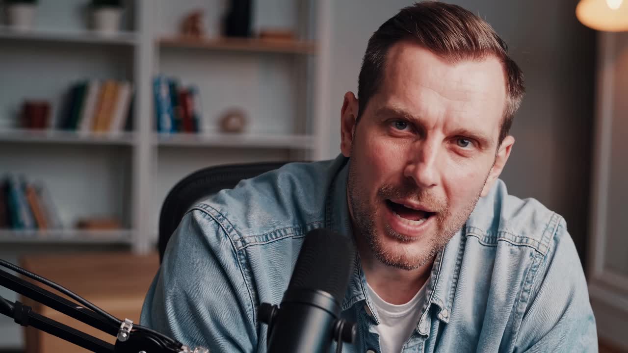 A smiling man in a denim shirt sits at a desk with a microphone, captured in a close-up angle