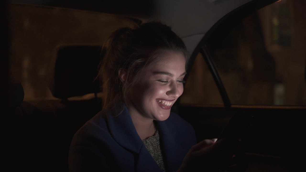 feliz niña sonriente montando en un taxi por la noche sentada en el asiento trasero y enviando mensajes de texto usando su teléfono inteligente. mujer feliz en el taxi