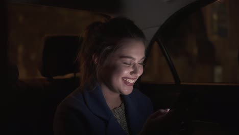 Una-Chica-Feliz-Y-Sonriente-Viajando-En-Un-Taxi-Por-La-Noche-Sentada-En-El-Asiento-Trasero-Y-Enviando-Mensajes-De-Texto-Usando-Su-Teléfono-Inteligente.-Mujer-Feliz-En-Taxi