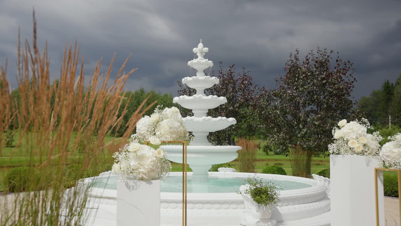 Elegant white wedding fountain surrounded by floral arrangements in a lush green garden with dramatic stormy clouds in the background