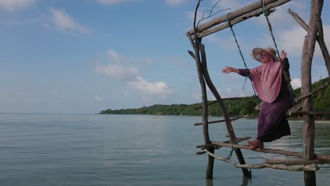 Fröhliche-Frau-Im-Sommerhut,-Die-Auf-Einer-Holzschaukel-über-Seichtem-Und-Ruhigem-Wasser-Auf-Der-Insel-Karimun-Jawa-Spielt