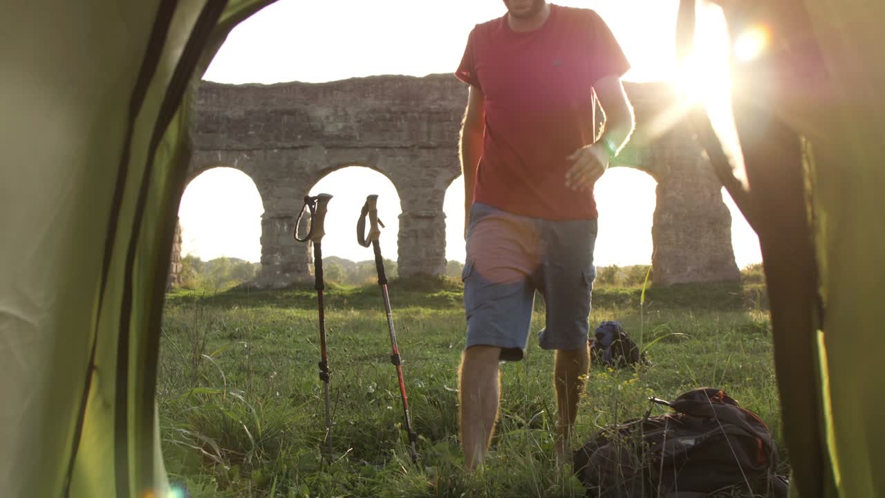 joven mochilero tocando la guitarra fuera de la tienda de campamento sentado en la hierba frente al acueducto romano arcos en el parco degli aquedotti ruinas del parque en roma al atardecer palos en el suelo cámara filmada dentro de la tienda pov cámara lenta