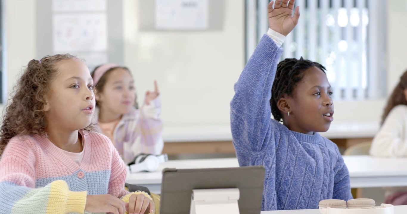School-age girls during lesson, blue-sweater girl raising then reaching then resting by tablet