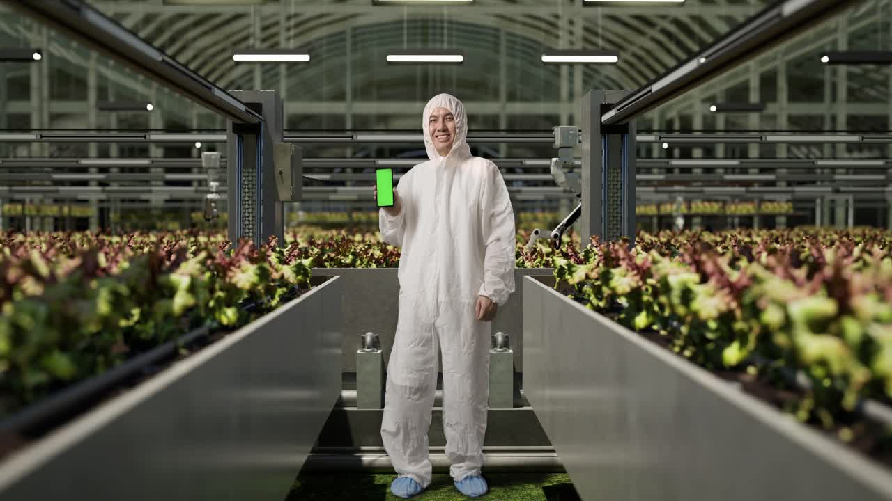 полное тело азиатского мужчины исследователь улыбается и показывает зеленый экран смартфона на камеру в то время как стоит в теплице с умными роботами-фермерами