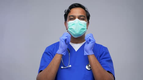 Concepto-De-Atención-Médica,-Profesión-Y-Medicina.-Médico-Indio-O-Enfermero-Con-Uniforme-Azul-Y-Mascarilla-Médica-Protectora-Que-Se-Pone-Guantes-Quirúrgicos-Sobre-Fondo-Gris.