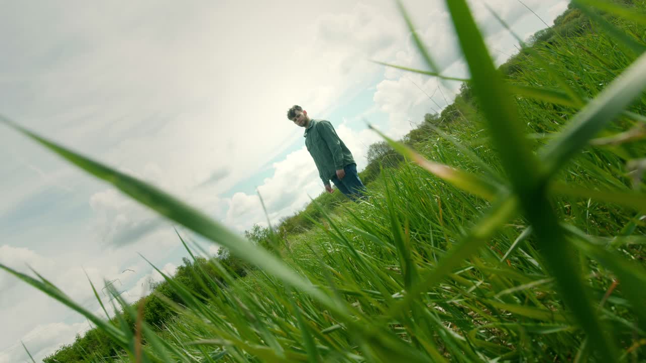 Low dutch angle view of man walking through grass with dramatic foreground blades, cinematic natural texture