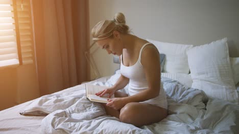 lovely young woman in white tank top and panties sitting on comfortable bed and reading