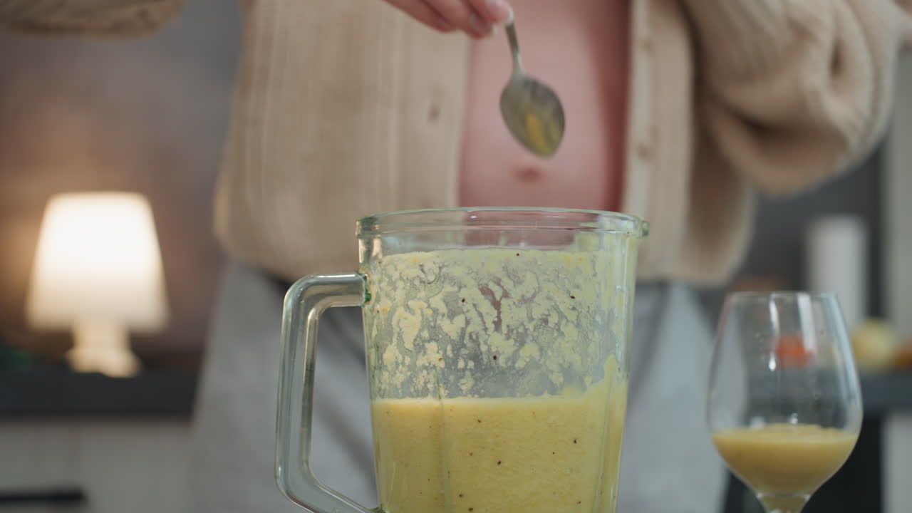 Donna incinta in cucina che prepara un frullato, accogliente scena mattutina con la pancia scoperta, cardigan morbido, caraffa del frullatore piena di purea verde, mano che mescola, che versa in un bicchiere, calma preparazione di uno spuntino prenatale.