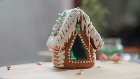 primer plano de una casa de pan de jengibre intrincadamente decorada con glaseado blanco, cuentas verdes y una ventana de azúcar verde brillante, bandeja de madera con salpicaduras doradas y fondo borroso