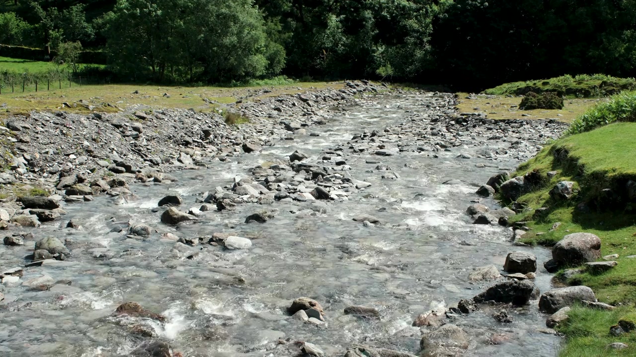 Toma panorámica estática de un río de montaña rocoso fluyendo a través de un tranquilo valle rural, con agua clara, rocas dispersas y exuberante bosque verde bordeando las orillas del río.