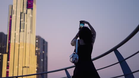 woman taking photo of illuminated skyscraper at dusk in city