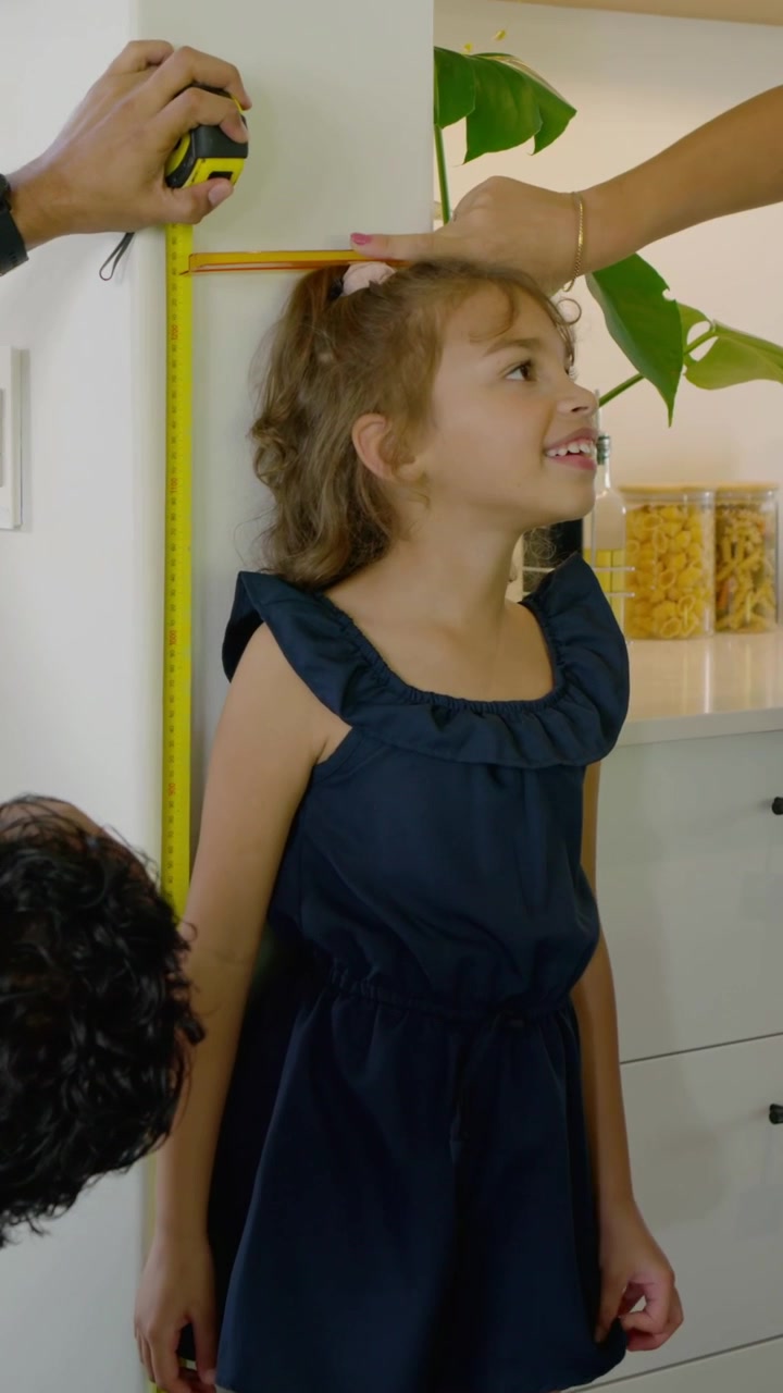Vertical video: Measuring parents tracking girl's growth in kitchen, placing book, holding tape