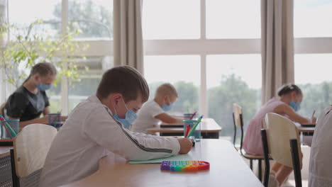 student in protective mask studying at school during corona virus pandemic. a masked teacher explains a new lesson topic to masked students. children write with pens in the classroom