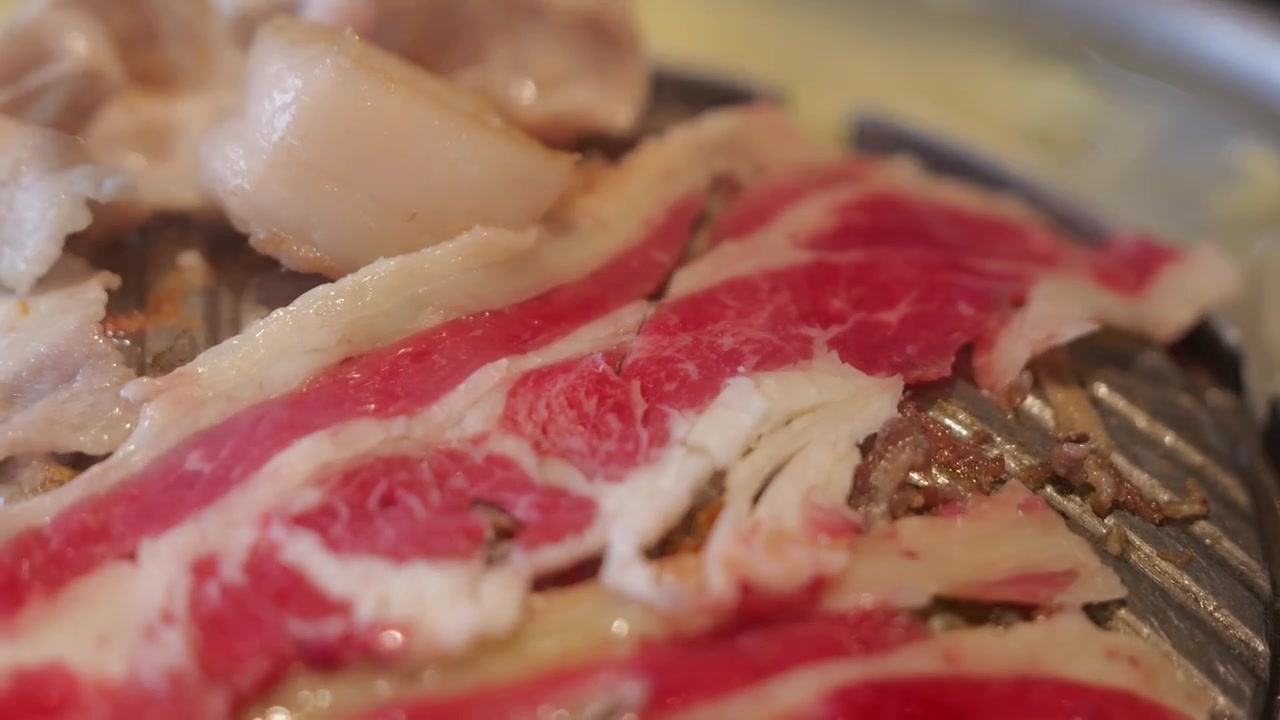 熱いタイ式バーベキュー鍋で焼かれる薄切り牛肉のクローズアップ。ジュージューと音を立てる肉、立ち上る煙、赤々と燃える熱、食欲をそそる焼き目を捉える。