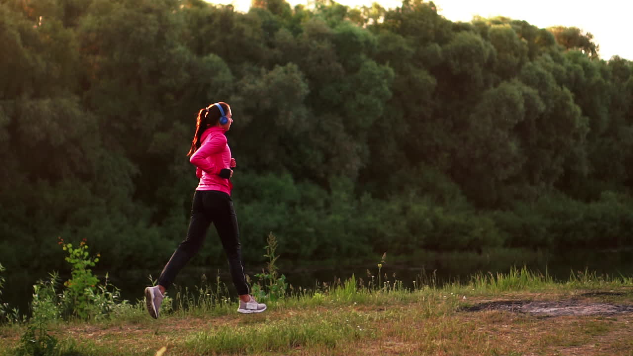 A girl in a pink jacket and black pants runs near the river in headphones preparing for the marathon