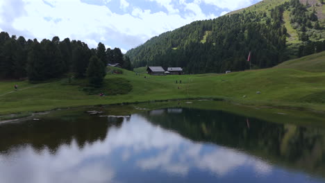 a serene mountain lake reflects the surrounding green hills, dense forest, and wooden cabins under a cloudy sky in a peaceful alpine setting