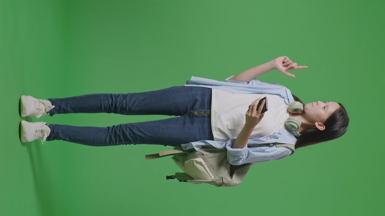 스마트폰을 사용하고 녹색 화면 배경 스튜디오에 서있는 동안 으로 가리키는 배을 가진 아시아 십대 소녀 학생의 전체 몸