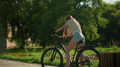 lady mounts bicycle near colorful garden, preparing for ride as she adjusts left foot on pedal, looking down with focus, surrounded by lush greenery, vibrant flowers, and tall trees