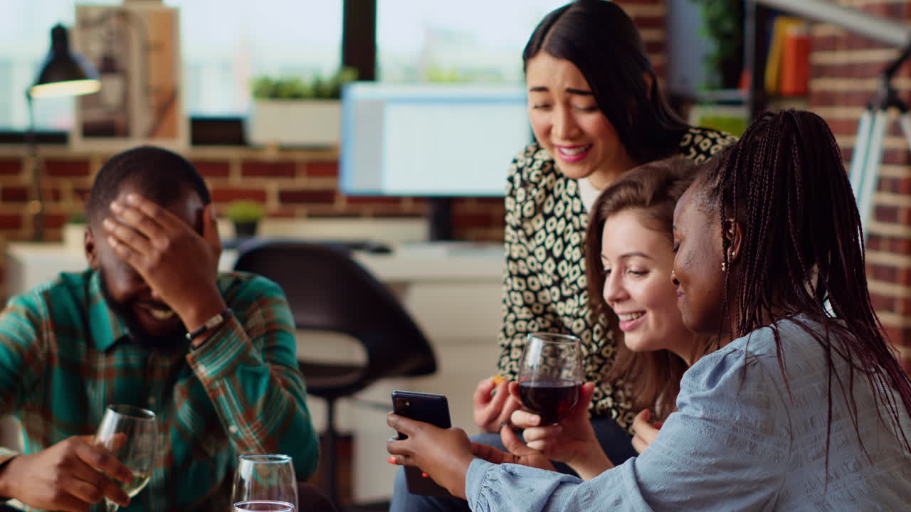 Joyful guests in living room watching internet clips on mobile phone while drinking wine