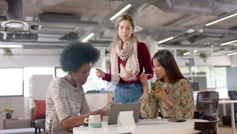 Diverse-female-creative-colleagues-discussing-using-tablet-and-eating-lunch-in-office,-slow-motion