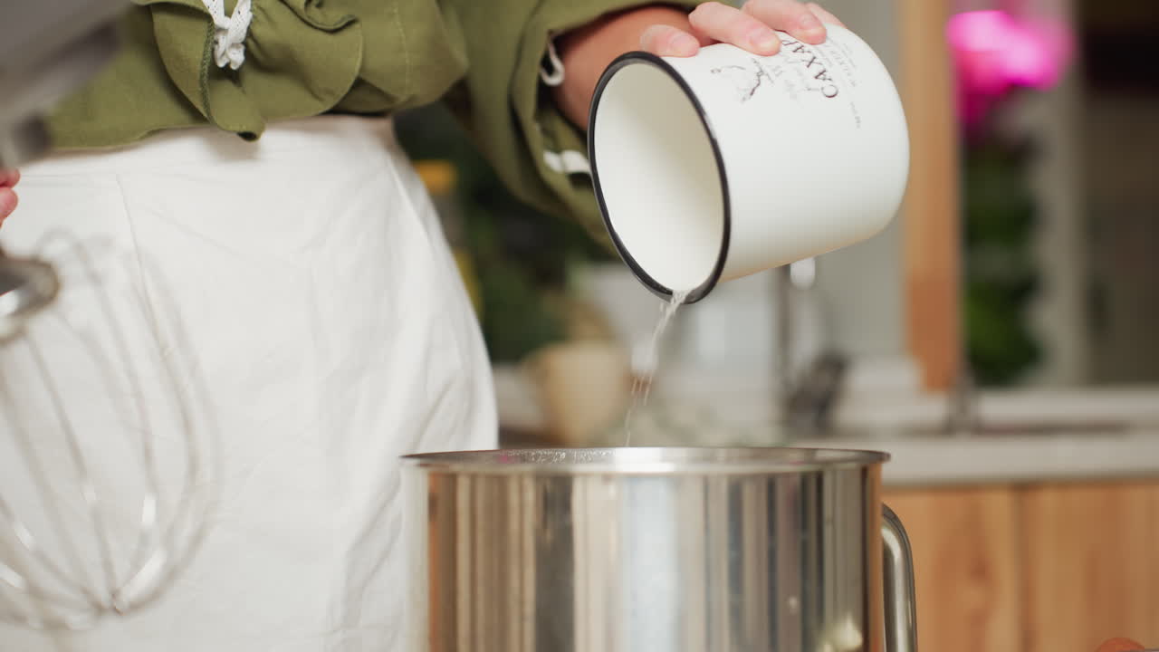 Close up of cook in green sleeve and white apron pouring sugar from vintage white cup into stainless bowl beside electric mixer during baking preparation in cozy home kitchen