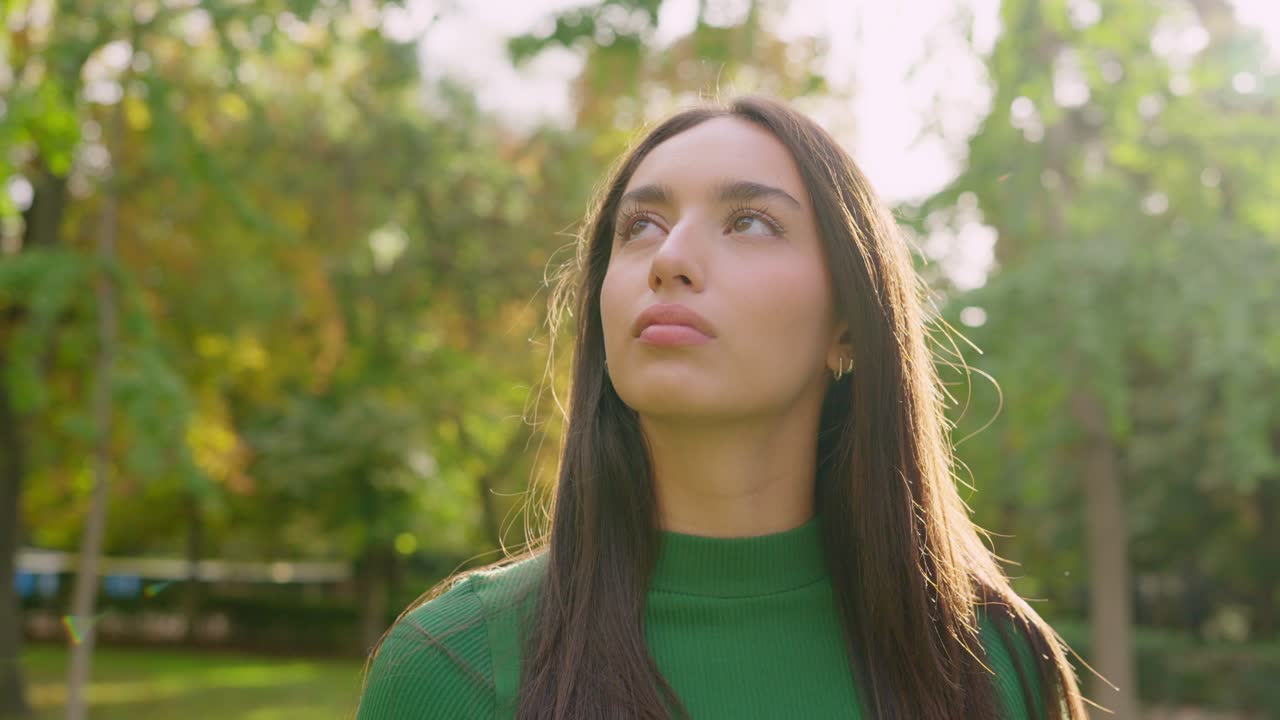 Thoughtful Young Woman in Green Shirt Looking Up in Sunny Park