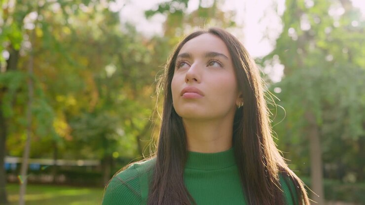 Thoughtful Young Woman in Green Shirt Looking Up in Sunny Park
