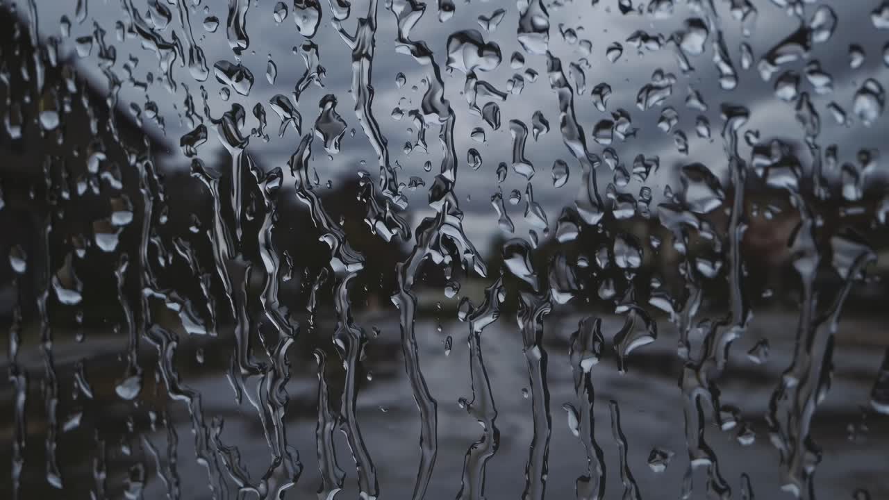Um vídeo em close-up captura gotas de chuva em uma janela, embaçando a vista de uma rua sombria e nublada.