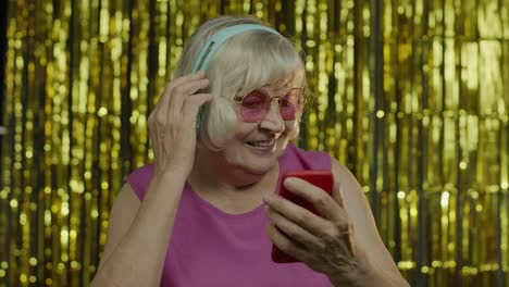 Stylish-happy-senior-old-woman-holding-cell-phone,-listening-to-music-in-headphones-and-dancing