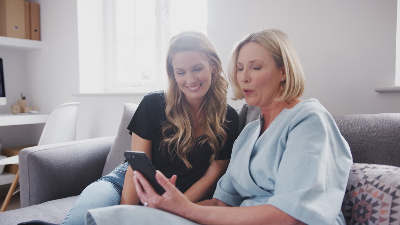 소파 에 앉아 휴대 전화 로 사진 을 보고 있는 성인 딸 과 함께 한 고령 어머니