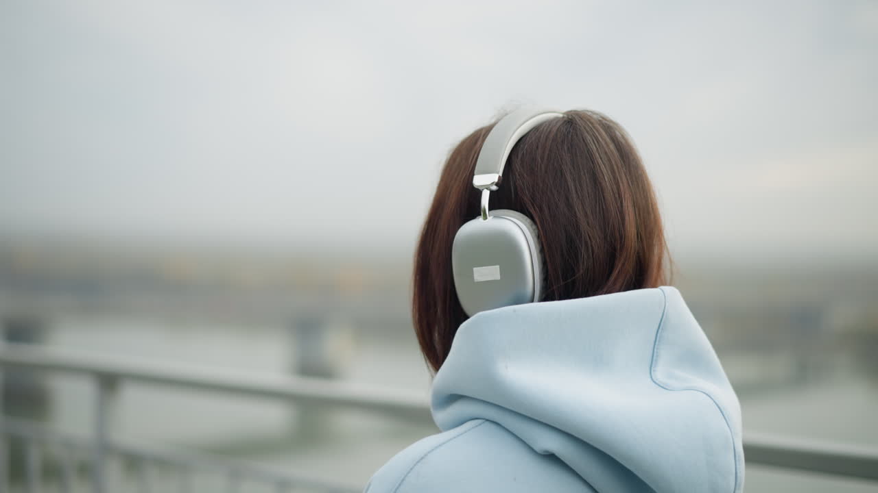 Backside view of woman walking and listening to music with headphones, blurred background featuring bridge, water, and iron rail, giving a calm and reflective atmosphere