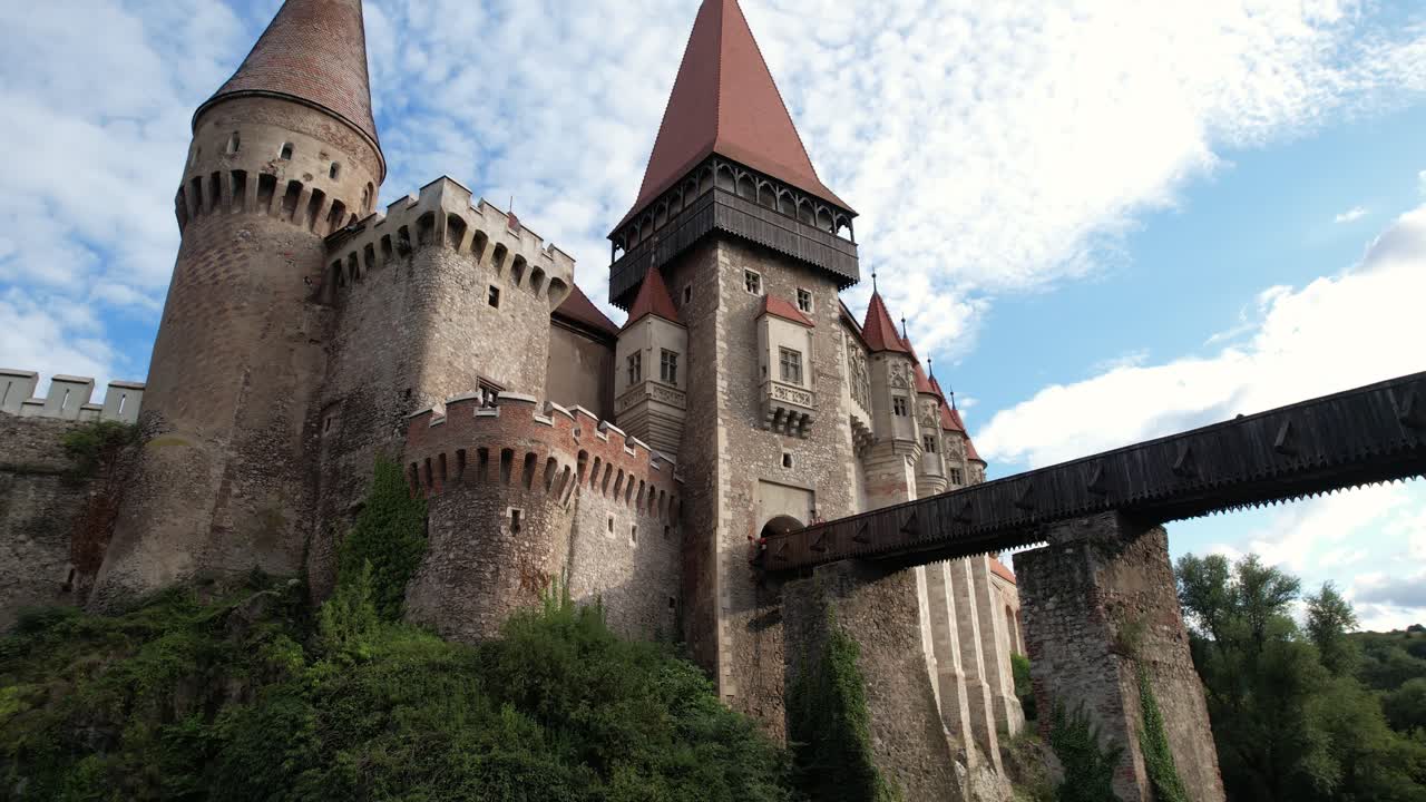 Low orbit drone shot of Corvin Castle, Romania. Historic Gothic fortress with scenic green hills under summer sky.