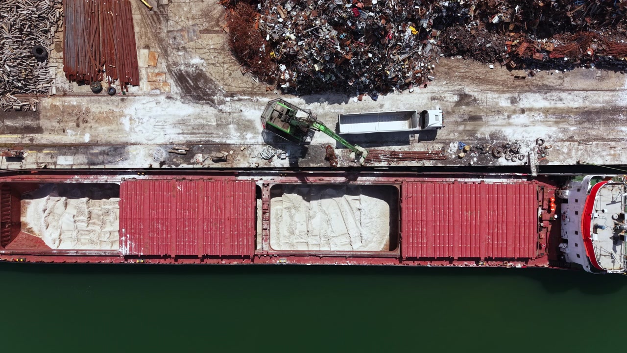 Grande nave da carico al molo mentre viene caricata di sabbia di giorno in una trafficata area industriale vicino a un porto con rottami metallici sullo sfondo.