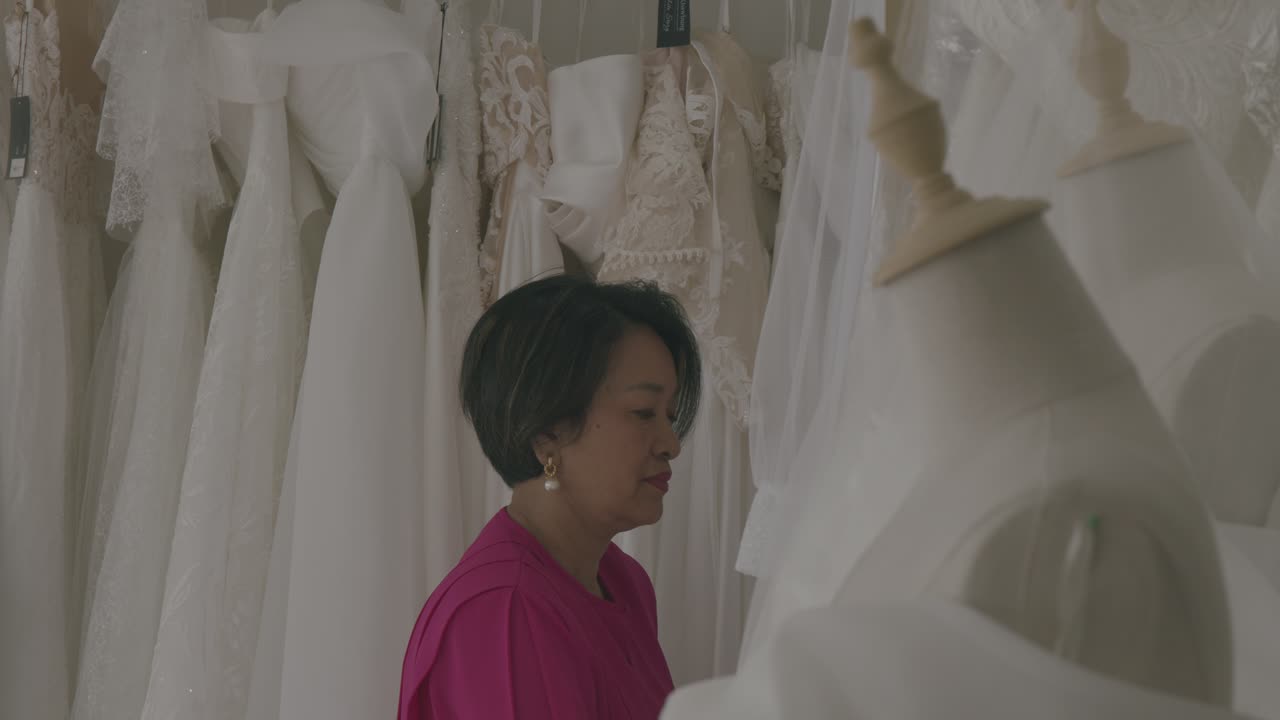 Inside a bridal store, a woman in pink examines luxurious wedding gowns on mannequins. Soft lighting enhances the serene ambiance, highlighting intricate lace and satin fabrics