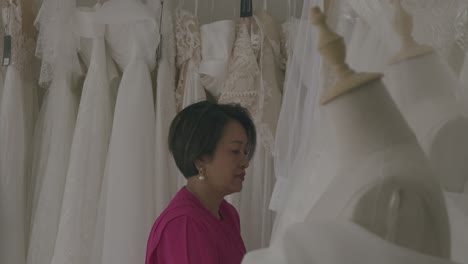inside a bridal store, a woman in pink examines luxurious wedding gowns on mannequins. soft lighting enhances the serene ambiance, highlighting intricate lace and satin fabrics