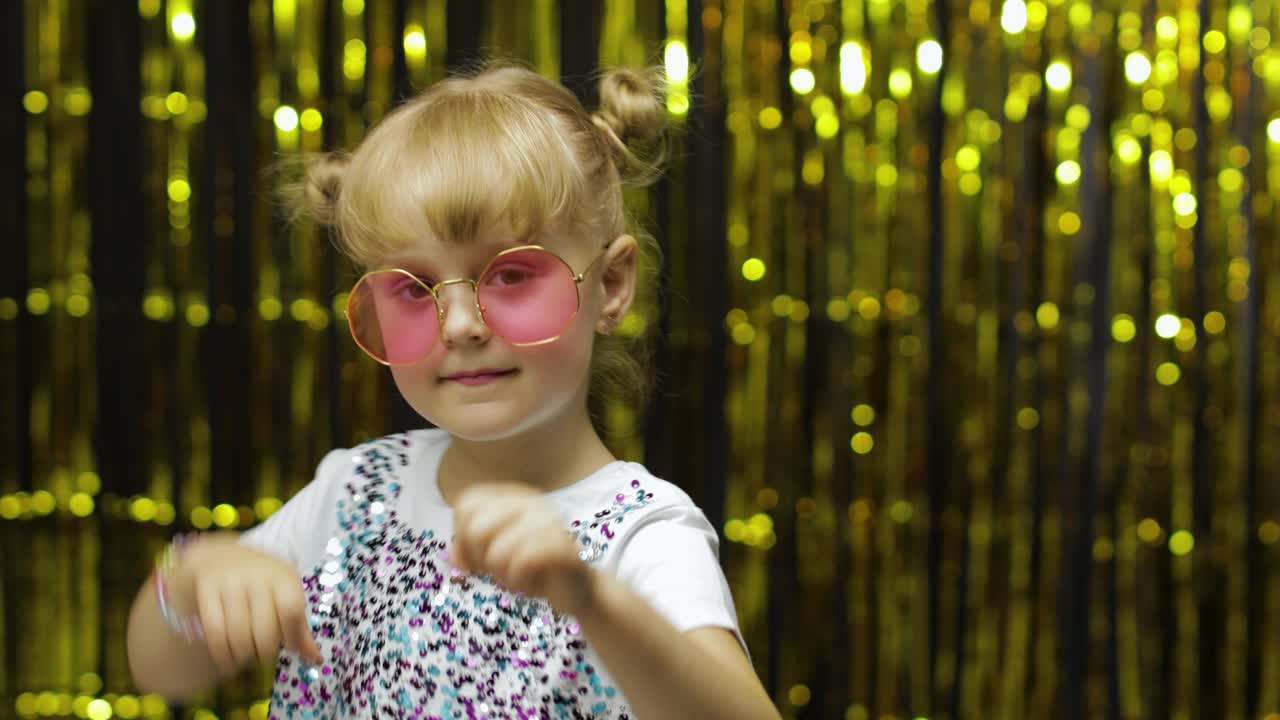 Child smiling, pointing fingers at camera. Girl posing on background with foil golden curtain